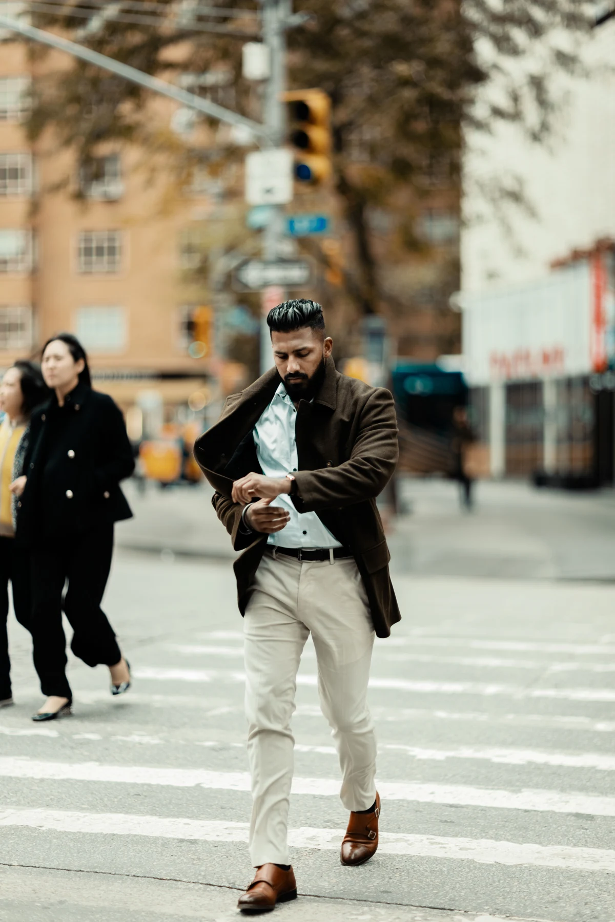 A person in a brown overcoat checking a watch while crossing a street.